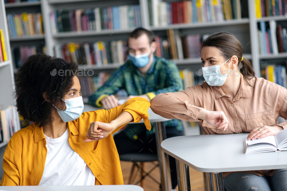 Two female student friends wearing protective face masks greeting each other bumping elbows while