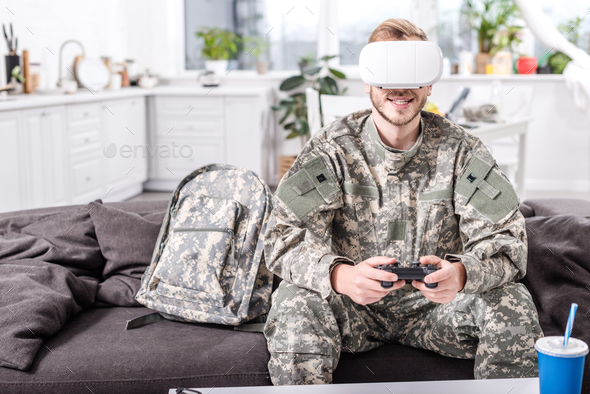 smiling army soldier in virtual reality headset playing video game on couch