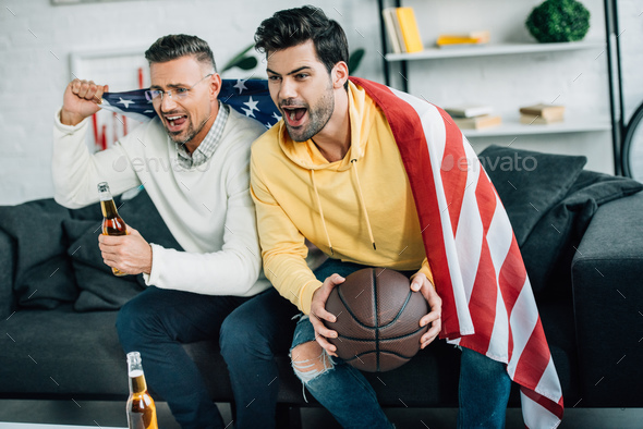 excited son and mature father wrapped in united states flag watching basketball game and screaming