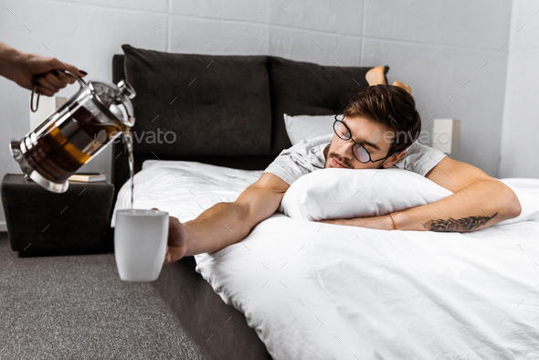 cropped shot of someone pouring tea from teapot while sleepy man in eyeglasses lying on bed and