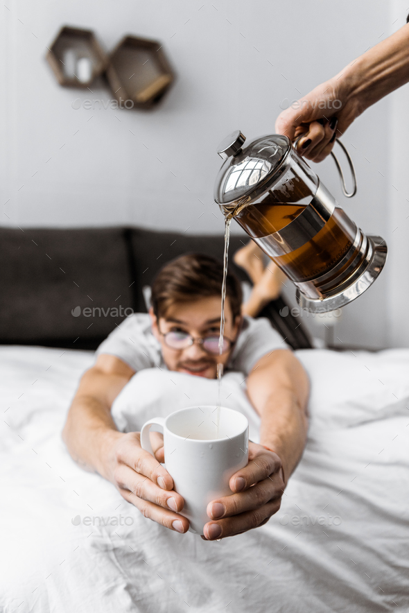 cropped shot of someone pouring tea to young man in eyeglasses lying on bed and holding cup
