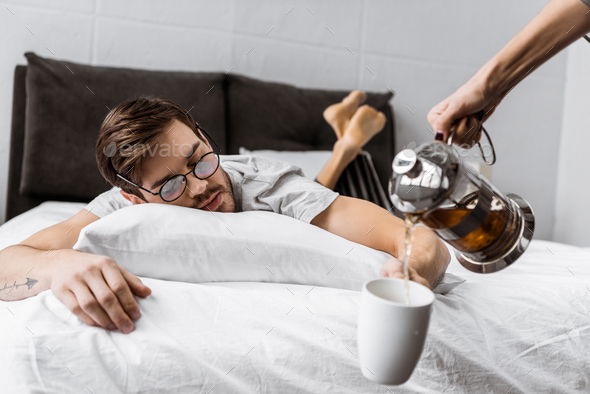 cropped shot of someone pouring tea into cup while sleepy man in eyeglasses lying on bed
