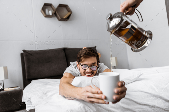 cropped shot of someone pouring tea to smiling man in eyeglasses lying on bed and holding cup
