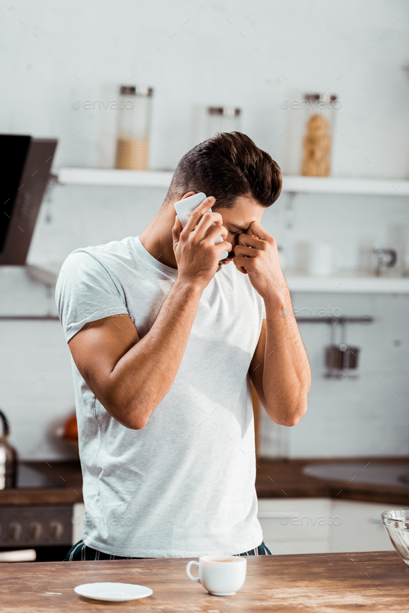 frustrated young man rubbing nose bridge and talking by smartphone at morning in kitchen