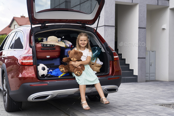 Portrait of caucasian girl holding a teddy bear and car trunk full of luggage in the background
