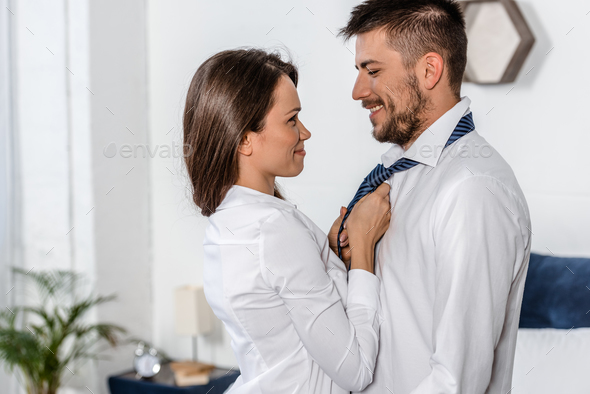 side view of cheerful girlfriend tying boyfriend tie in morning on weekday in bedroom, social role