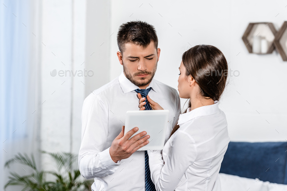 girlfriend tying boyfriend tie while he using tablet in morning at home, social role concept