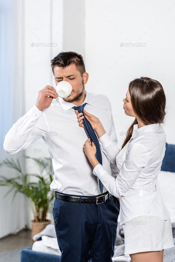 girlfriend tying boyfriend tie while he drinking coffee in morning at home, social role concept