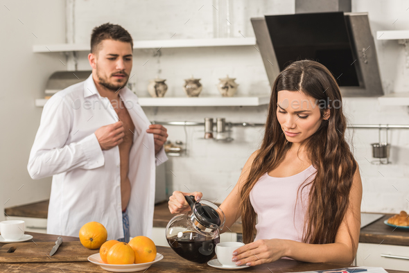 boyfriend buttoning shirt and girlfriend pouring coffee into cup at home, social roles concept