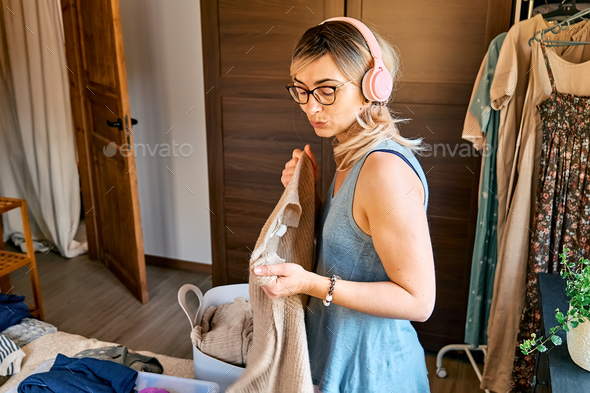 Blond woman in headphones listening music, dancing and singing while making order of clothes.