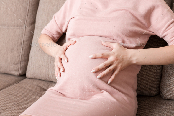 Pregnant Lady Having Massaging Lower belly Sitting On Sofa Indoor. Pregnancy Problems Concept.