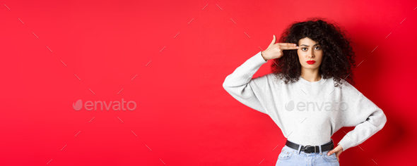 Annoyed caucasian woman with curly hair, showing hand gun sign on head, blow her mind off, standing