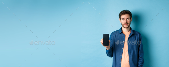 Annoyed young guy showing empty smartphone screen and roll eyes up bothered, standing on blue