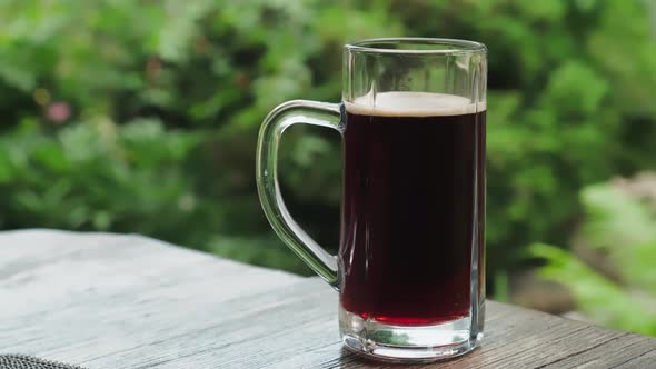 A glass of cold frothy dark beer on the table against the backdrop of nature.