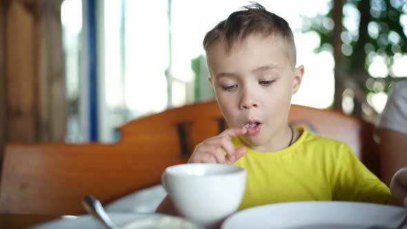 Little Boy Is Sitting in a Cafe Eating Pancakes with Sour Cream and Drinking Tea alt