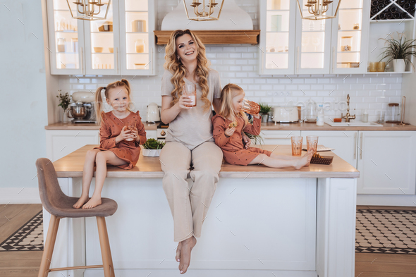 Pregnant Swedish mom sitting on kitchen table with little daughters dressed in beige casual clothes