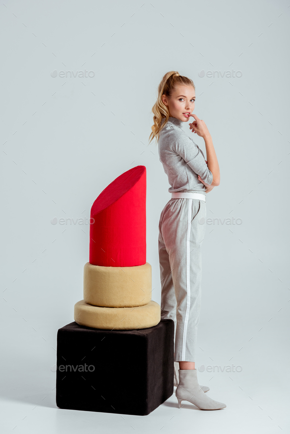 beautiful woman biting finger and posing near big red lipstick model on grey background