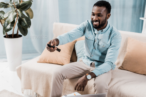 smiling african american man sitting on couch and using remote controller