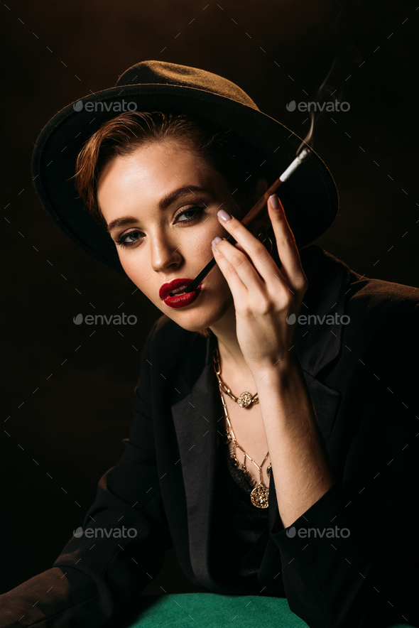 attractive girl in jacket and hat smoking cigarette at table isolated ...