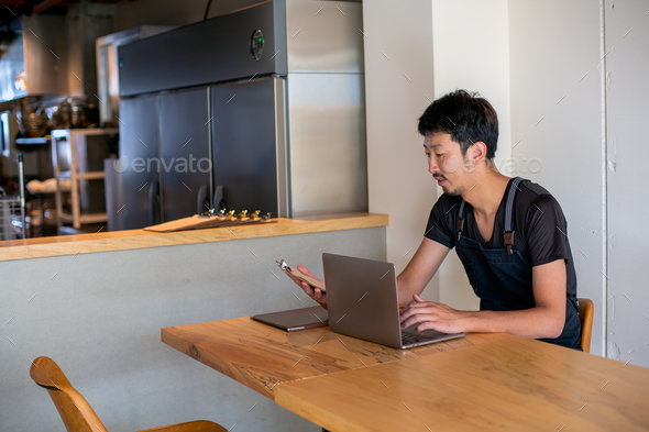 A man seated at a table using a laptop computer, owner and manager of a small restaurant. - Stock Photo - Images