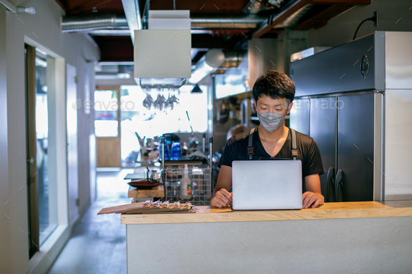 A man in a face mask in a restaurant kitchen, using a laptop, the owner or manager.