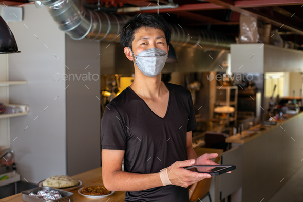 A man working in a restaurant, wearing a face mask, by an open kitchen, holding a digital tablet. - Stock Photo - Images