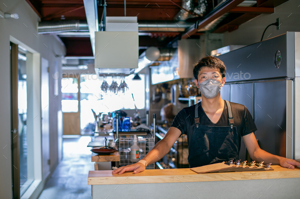 A man in a face mask at the counter of a restaurant kitchen, the owner or manager.