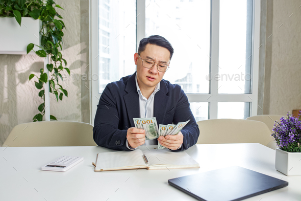 Dissatisfied asian entrepreneur holding cash US dollars, frustrated by low income. Low salary.