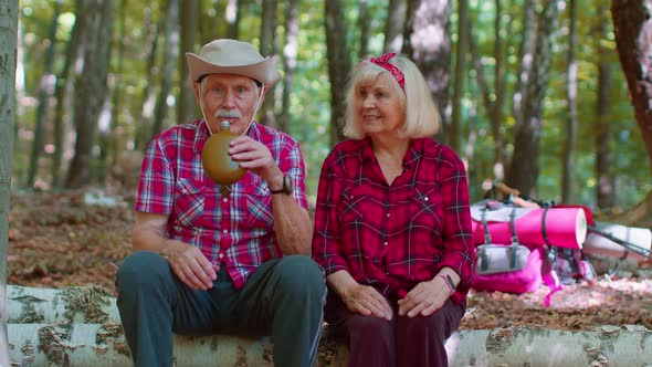 Tired Thirsty Senior Hikers Grandmother Grandfather with Backpacks Relaxing Drinking Water in Wood alt