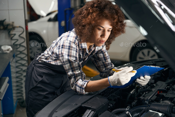 Female technician in work clothes stands near car with open hood