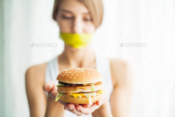 Young woman with duct tape over her mouth, preventing her to eat junk food. Healthy eating concept