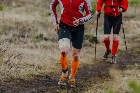 two runners run muddy trail sports race, knee is covered in blood injury  from fall Stock Photo by realsportsphotos