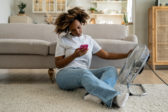 Young black woman using mobile phone while relaxing in front of electric fan at home