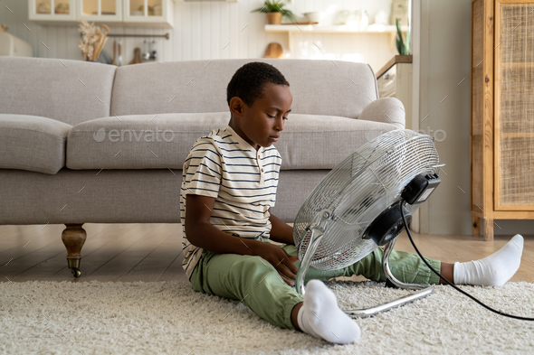 Curious child African American boy sitting on floor at home touching electric floor fan. Summertime