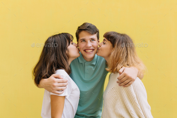 Two teen girls kissing handsome boystanding between them - isolated on yellow. - Stock Photo - Images