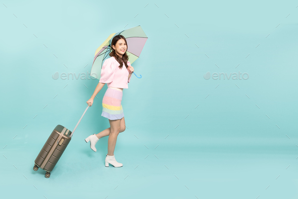 Young Asian woman traveler drag luggage and holding pastel umbrella isolated on green background