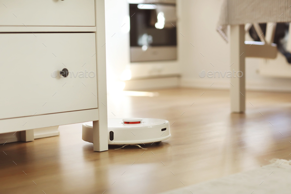 White robotic vacuum cleaner on laminate floor cleaning dust in living room interior. Smart elect