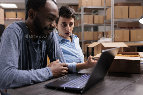 Diverse employees sitting at table in warehouse checking transportation logistics on laptop computer