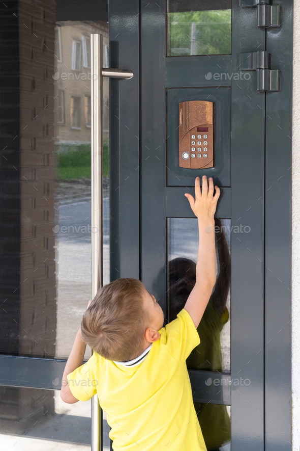 A little boy reaches for the bell to ring the doorbell, view from the back. A child standing near