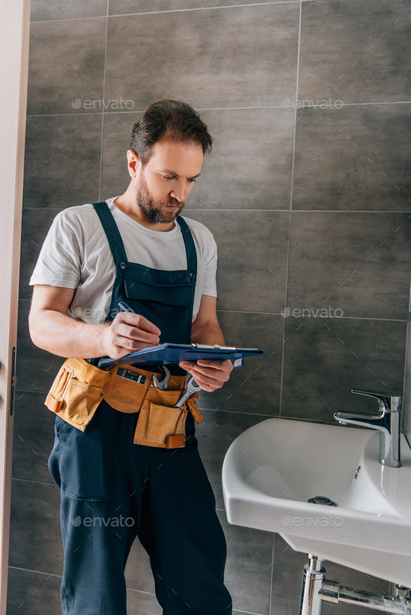 handsome male plumber with toolbelt writing in clipboard near broken sink in bathroom