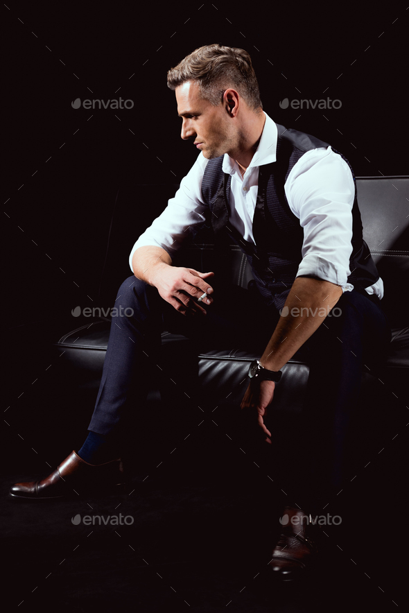 handsome man in formal wear sitting on couch with cigarette isolated on black