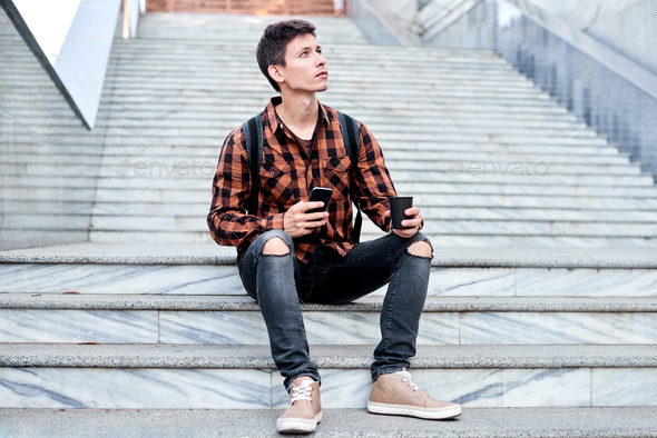 stylish guy with mobile phone and coffee sitting on granite stair. student on coffee break