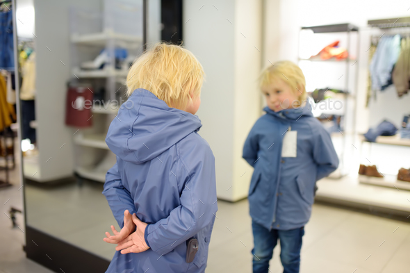 Cute little boy is trying on a new coat in front of a mirror in a store or shopping mall.