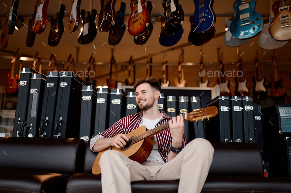 Happy man musician playing guitar over string instrument shop store background