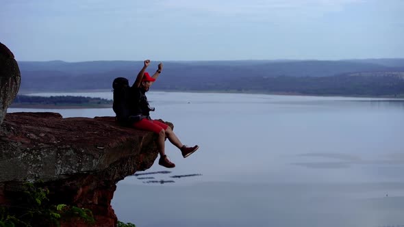 slow-motion of cheerful hiker man sitting and gesture raised arms on the edge of cliff alt