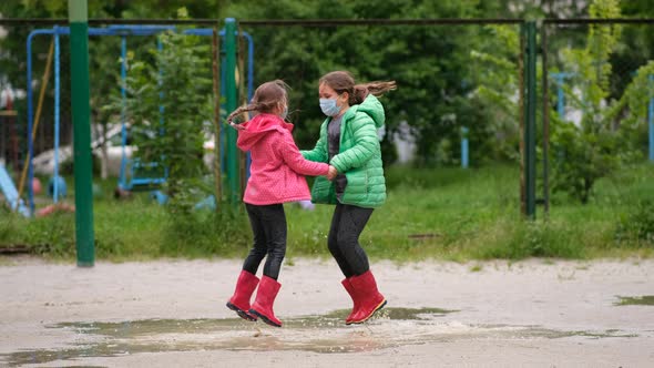 Two little girls in medical masks and rubber boots jump through puddles on a spring day after rain