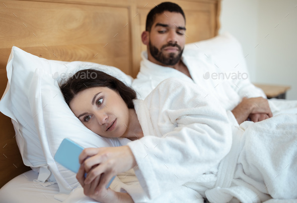 Husband Looking At Cheating Wife Texting On Smartphone In Bedroom