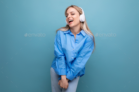 shy blonde girl in a casual shirt listens to songs in big headphones and sings along on a blue
