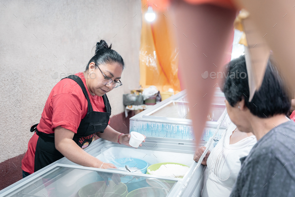 An Hispanic adult woman is serving an ice cream on a cup in a street market stall