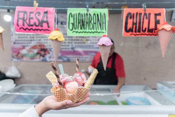 A hand is holding a traditional Mexican decorated ice cream in a street market with flavor signs